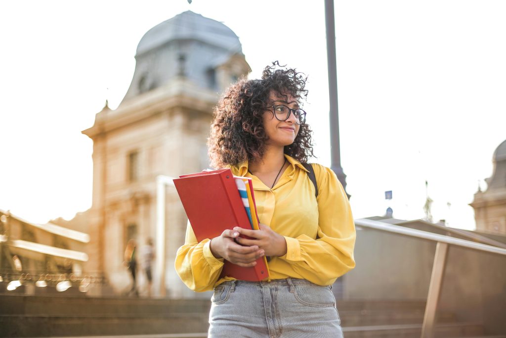 Student Area Smiling female student with curly hair and glasses holding books on campus steps.